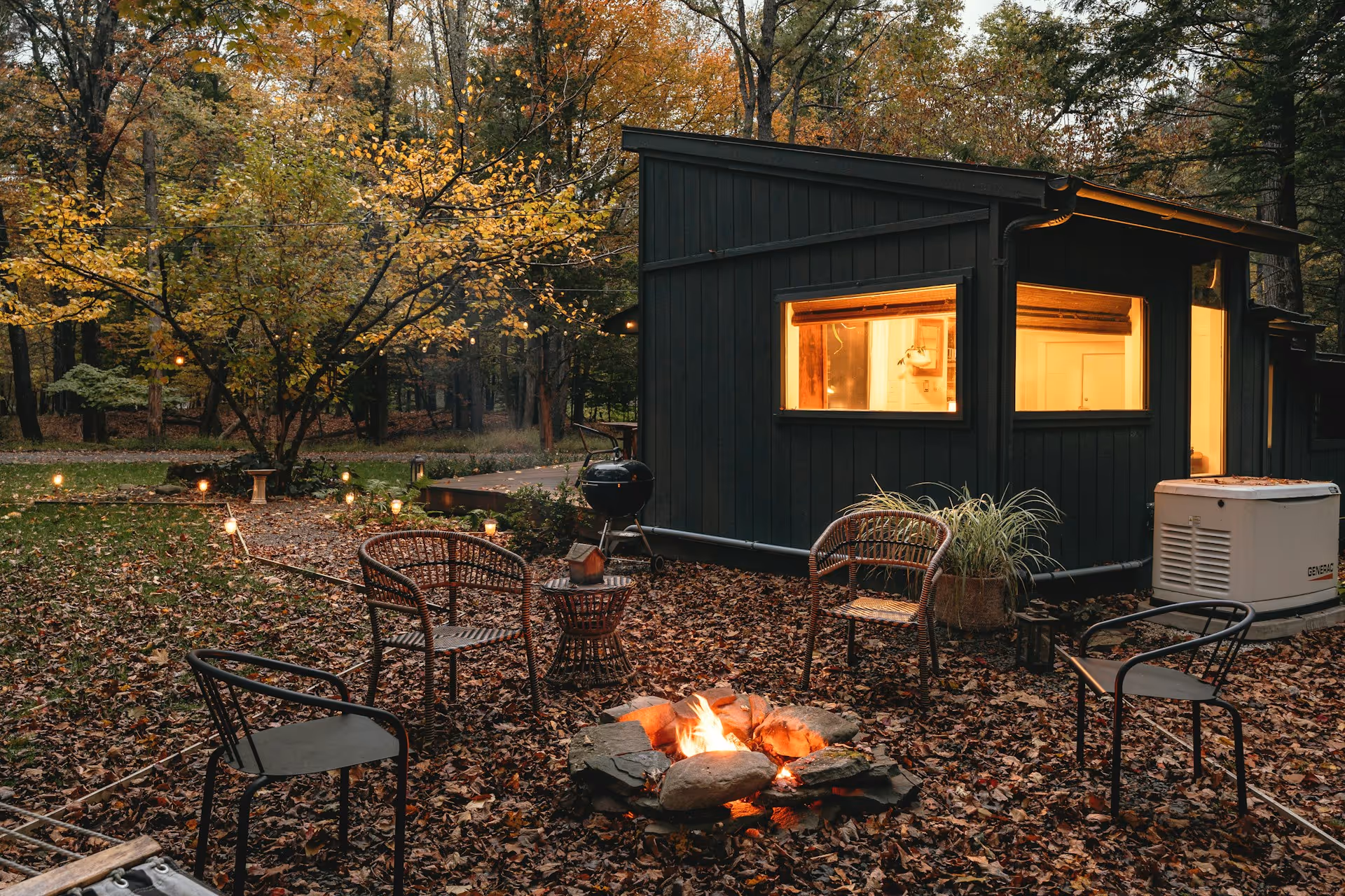 Outdoor firepit surrounded by four chairs on autumn leaves next to a dark cabin with warm interior lighting.