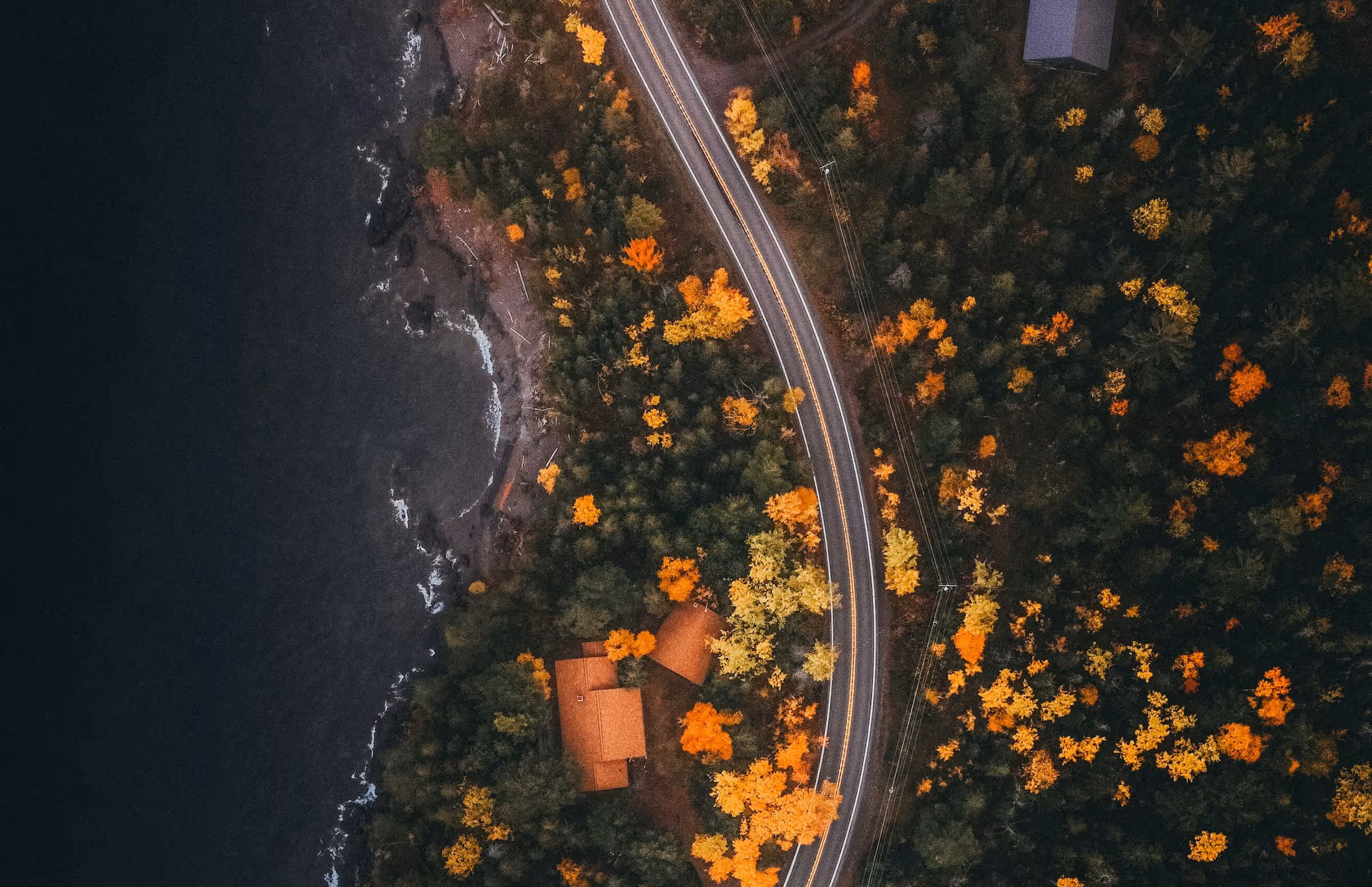 Aerial view of a winding road through a forest with autumn foliage next to a dark body of water and an orange-roofed house.