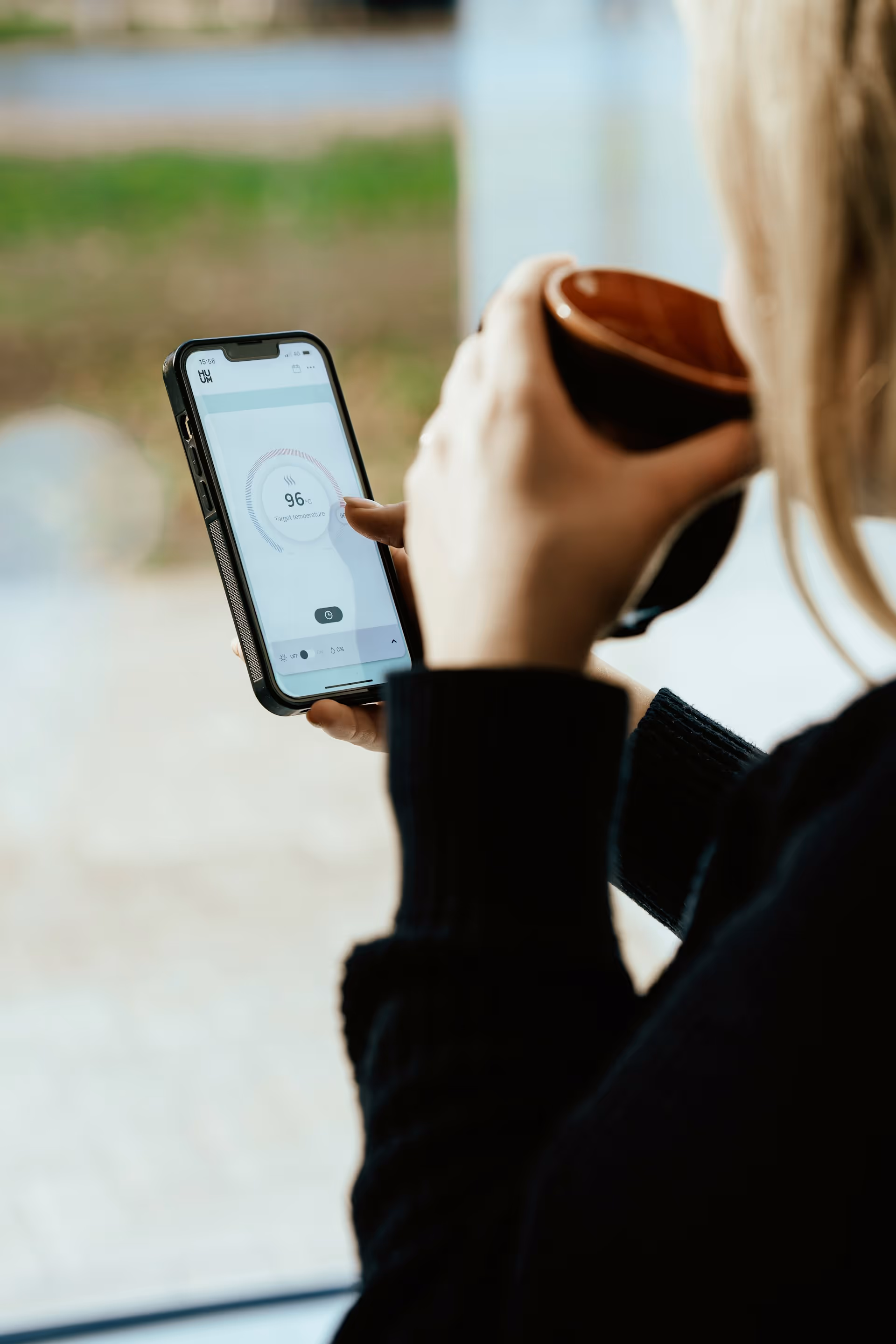 Person holding a smartphone displaying a temperature control app set to 96°C while holding a brown cup.